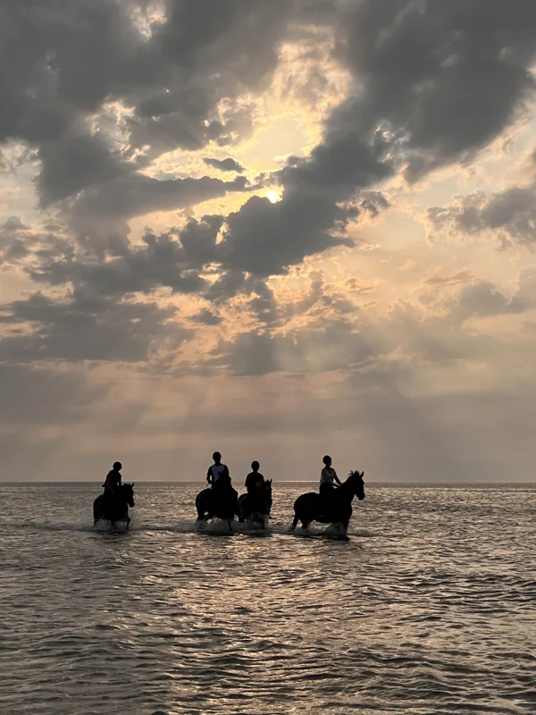 Four riders in shallow water beneath sunlight breaking through clouds